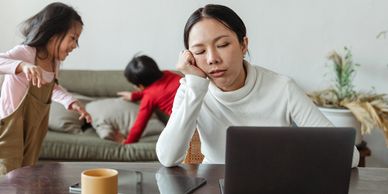 A woman of Asian decent seated in front of a computer in a home while two children play behind her