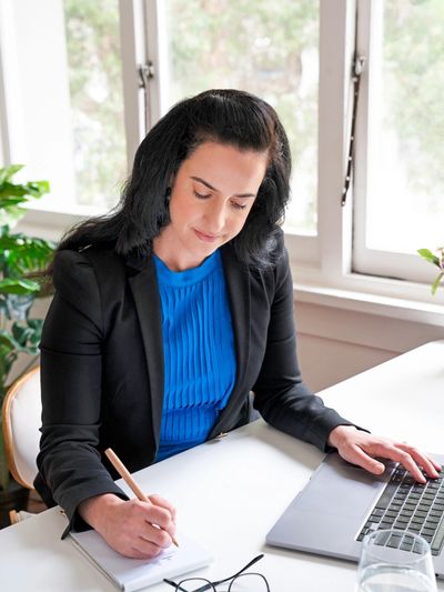 Focused woman in blue blouse and black blazer writing notes at her desk with a laptop.