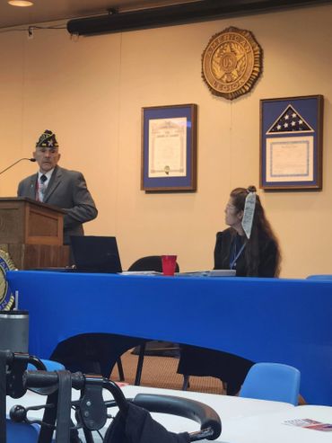 A man in military attire speaks at a podium in a formal setting with a woman seated at a table.