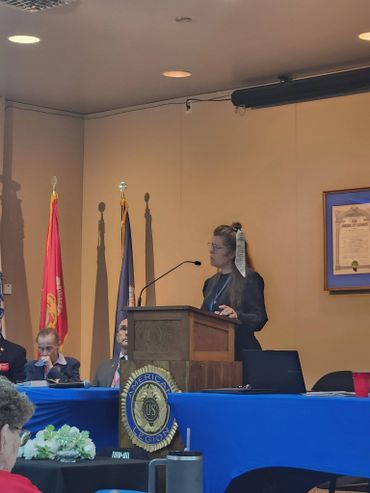 A woman speaking at a podium in a formal setting with flags behind her.