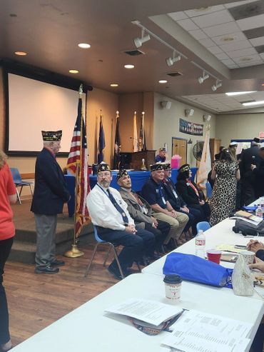 Veterans in uniform seated during a ceremony with flags in the background.