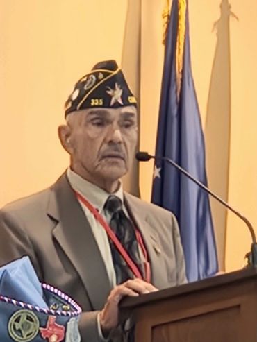 Veteran in uniform speaking at a podium with flags behind.
