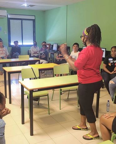 A woman speaks to a seated group in a classroom with green walls.