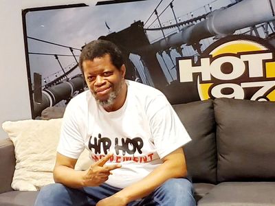 A man in a white shirt sitting on a couch with a Hot 97 backdrop.