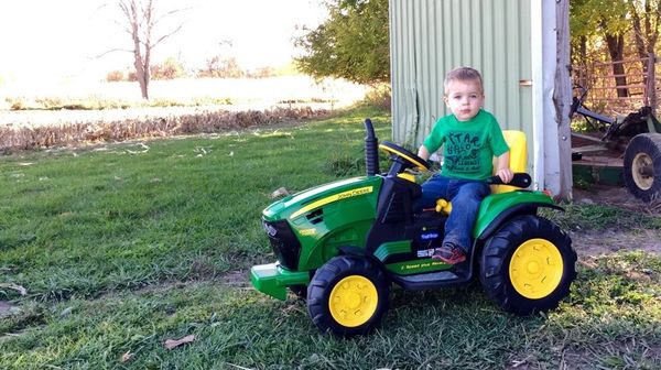 A young boy riding a green toy tractor outdoors near a barn.