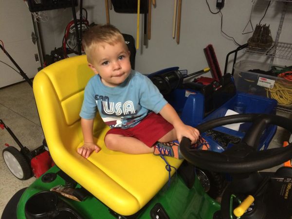 A young boy sits on a yellow seat of a green lawn mower indoors.
