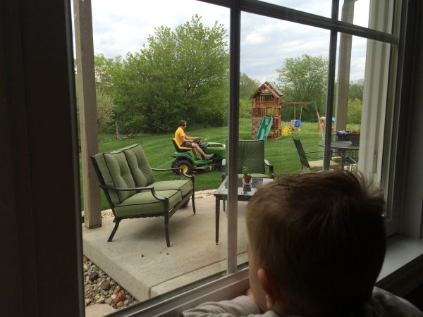 Child watches a person mowing the lawn from inside the house.