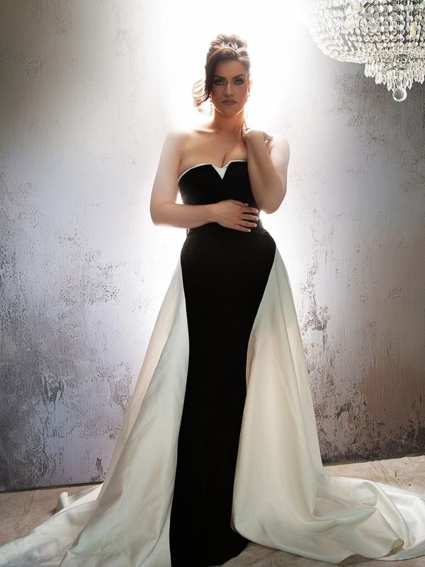 Woman in elegant black and white gown stands under chandelier with dramatic lighting.