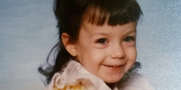 A smiling toddler girl with bangs and a frilly yellow dress.