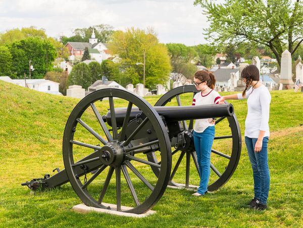 Two girls examine a Civil War cannon on a grassy hill overlooking a town.