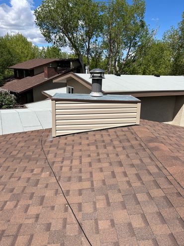 Rooftop chimney vent with brown shingles and nearby houses under a blue sky.