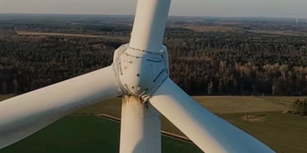 Close-up of a wind turbine with rust near the hub against a forest and field background.