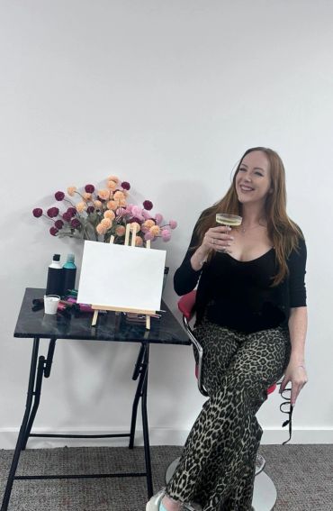 Woman smiling with a drink beside a blank canvas and colorful flowers.