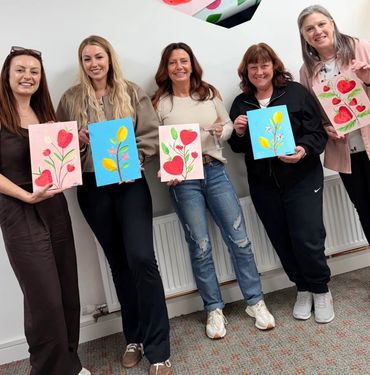 Five women proudly display their colorful floral paintings indoors.