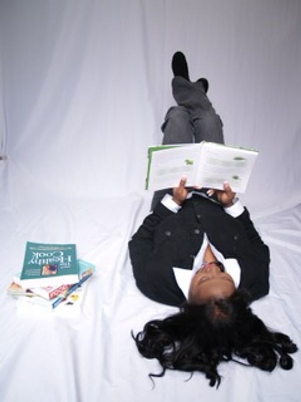 A woman lies on her back reading a book with legs raised, surrounded by a few books.