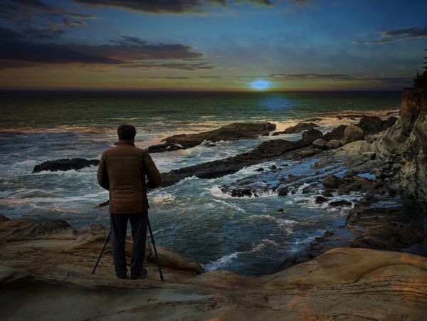 A person stands with a tripod on a rocky shore watching the sunset over the ocean.