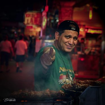 Smiling street vendor pointing at the camera in a vibrant night market.