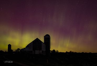 Northern lights glow above a rural barn and silo at night.