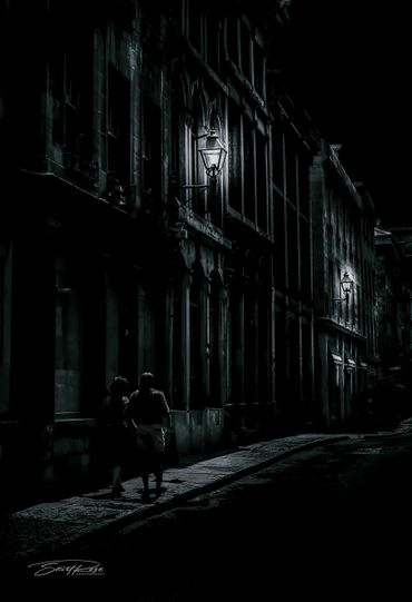 Two people walk on a dimly lit street at night under old-fashioned lamps.
