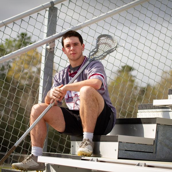 A young man in lacrosse gear sitting on bleachers holding a lacrosse stick.