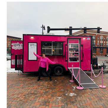 A smiling woman poses in front of a bright pink food truck on a city street.