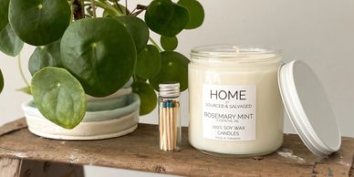 A rosemary mint scented candle and matches on a wooden shelf with a green plant.