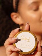 Hand holding a golden compact makeup product with blurred face background.