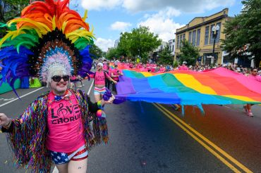 Person in colorful headdress holding a rainbow flag at a parade.