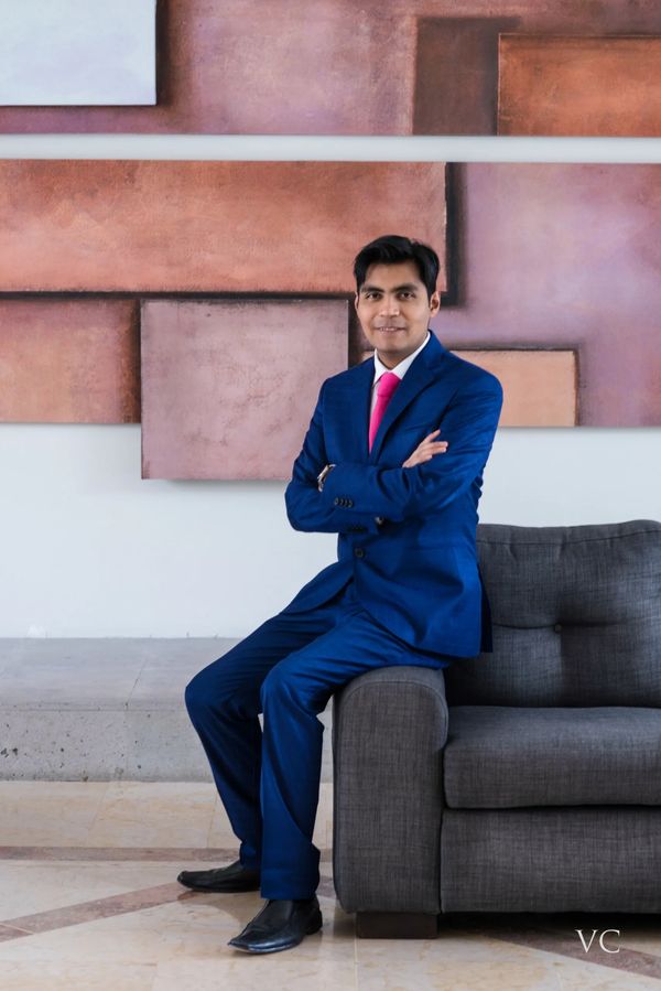 Man in blue suit with pink tie sitting on a gray couch, smiling confidently.