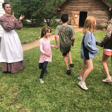 Children holding hands and playing outdoors with a woman in historical attire clapping.