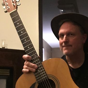 Man holding an acoustic guitar, wearing a hat indoors.