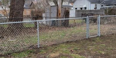 A rusty chain-link fence encloses a patchy backyard with leafless trees and neighboring houses.
