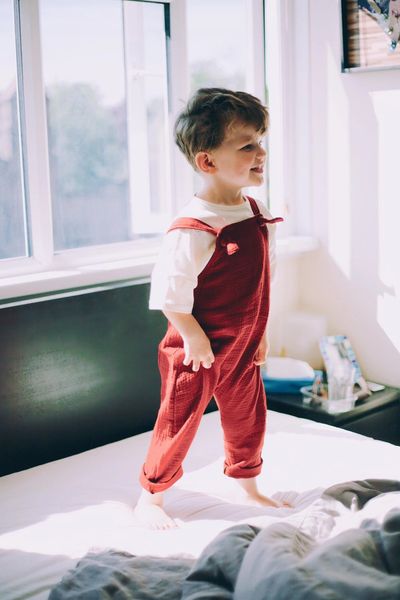 a boy standing on a bed holding his maroon coloured dungarees