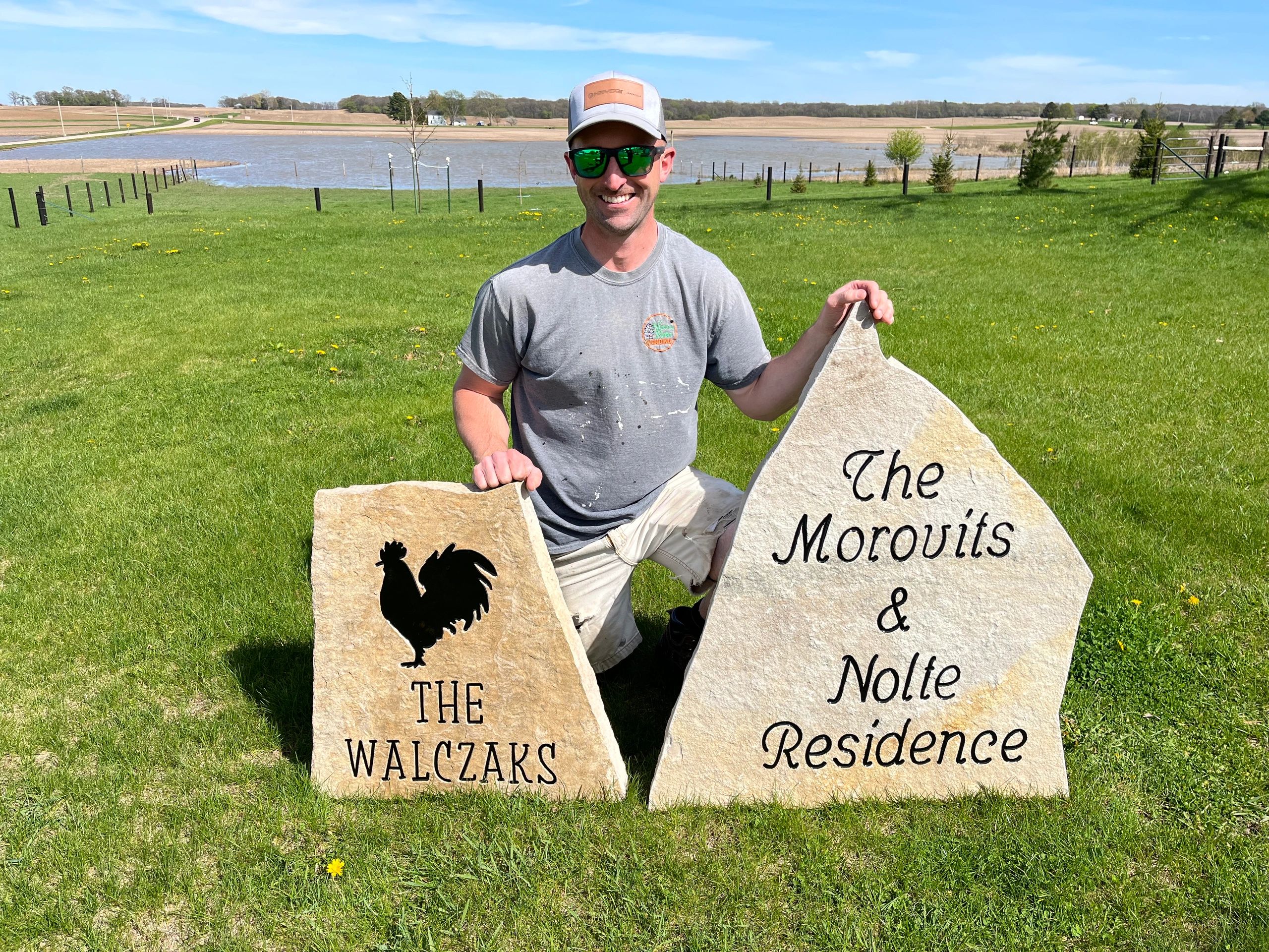 Man kneeling on grass holding two personalized stone signs on a sunny day.
