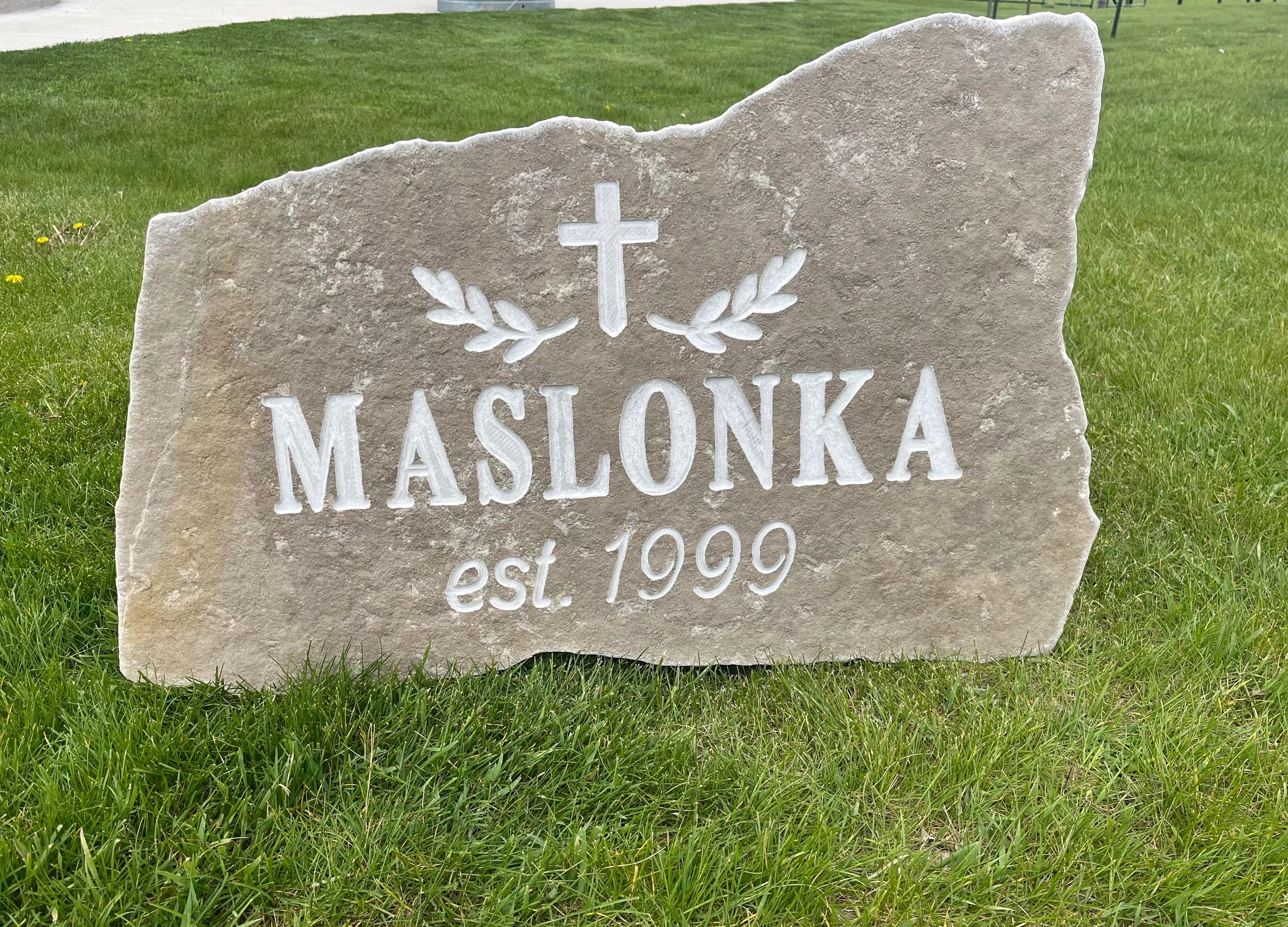 Stone sign with cross and leaves engraving, reads "MASLONKA est. 1999".