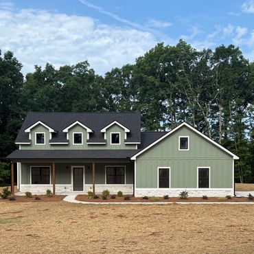 Modern green house with black roof and dormer windows surrounded by trees.