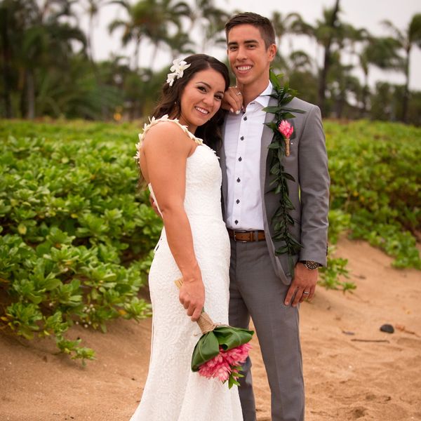 A happy couple in wedding attire posing on a beach with greenery in the background.