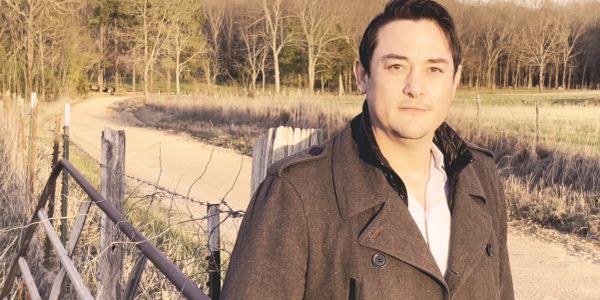 Man standing outdoors near a fence and a dirt road.
