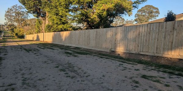 A long wooden fence along a dirt pathway with trees and houses in the background.