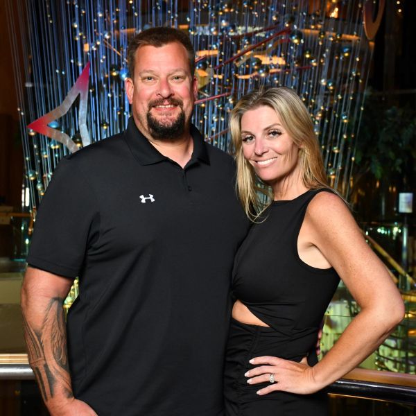 A man and woman pose together, both smiling, in a stylish indoor setting with decorative lights.