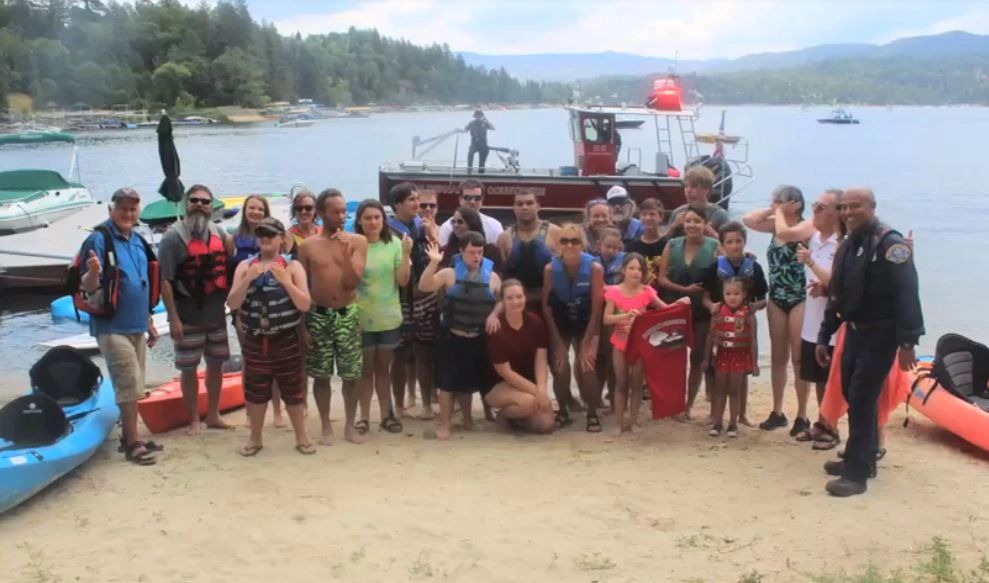 Group of people posing on a lakeside beach with kayaks and a rescue boat in background.