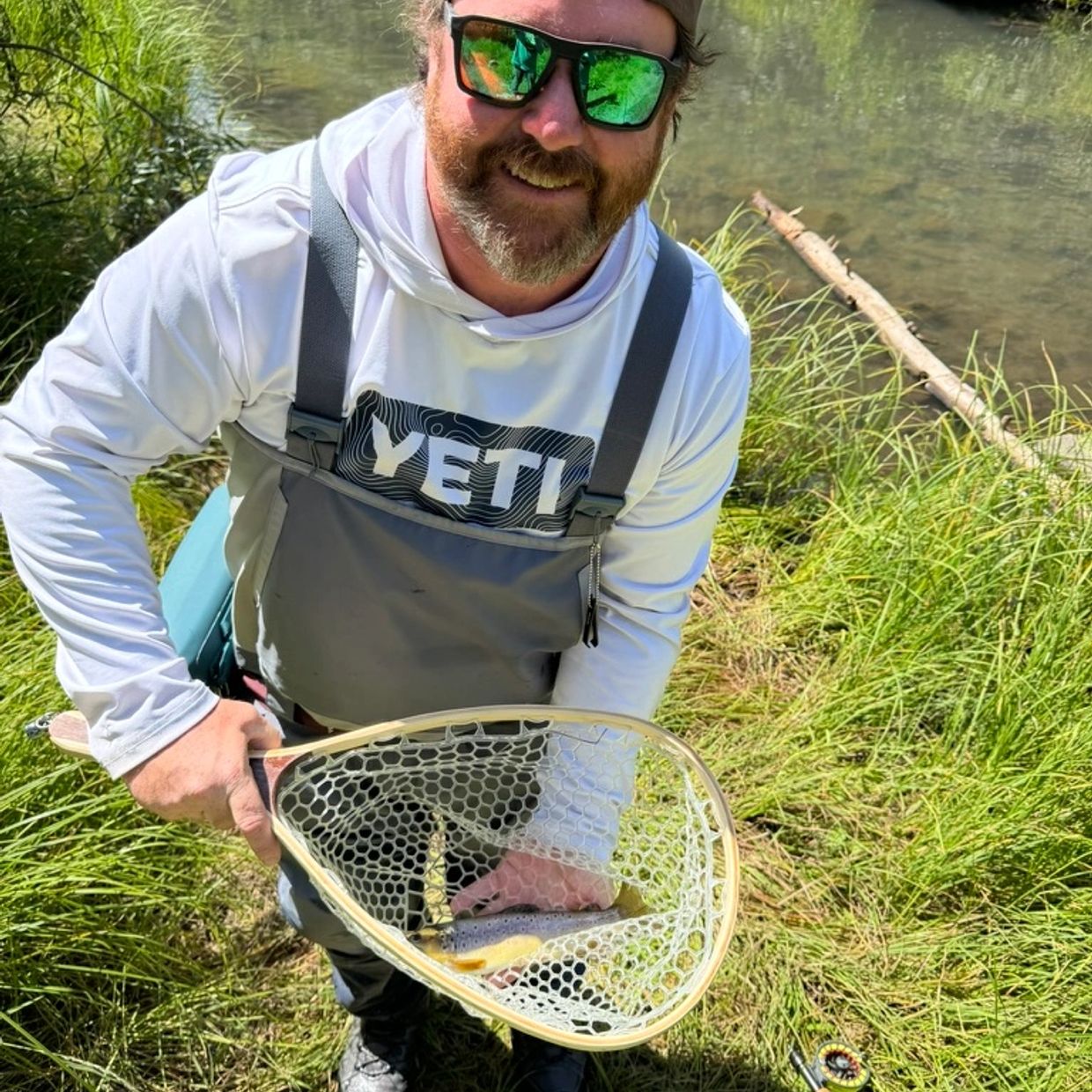 Man holding a fish in a net by a river, smiling at the camera.