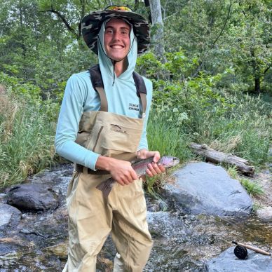 Smiling man in fishing gear holding a rainbow trout in a forest stream.

