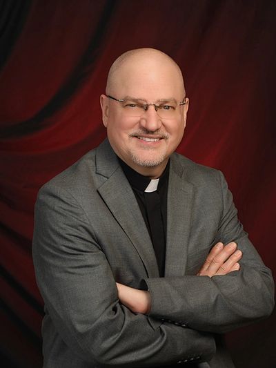 Smiling clergyman in gray suit with glasses crosses arms against a red backdrop.