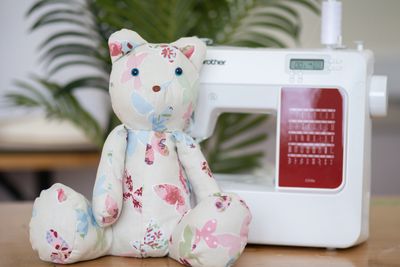 A floral fabric teddy bear sits next to a white sewing machine.