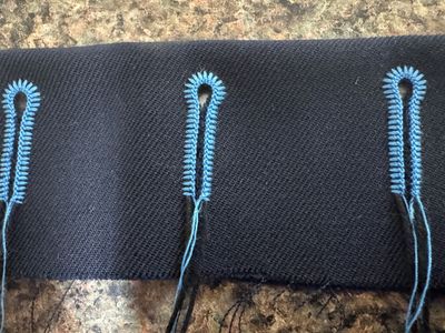 Black fabric with blue embroidered buttonholes in progress.