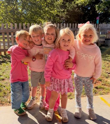 Five children smiling outside near a fence.
