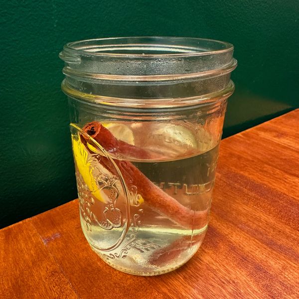 Mason jar with clear liquid, cinnamon stick, and lemon slice on wooden table.