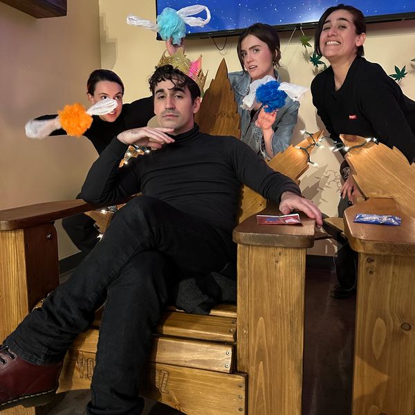 A man wearing a crown sits on a large wooden chair with three women playfully posing behind him.