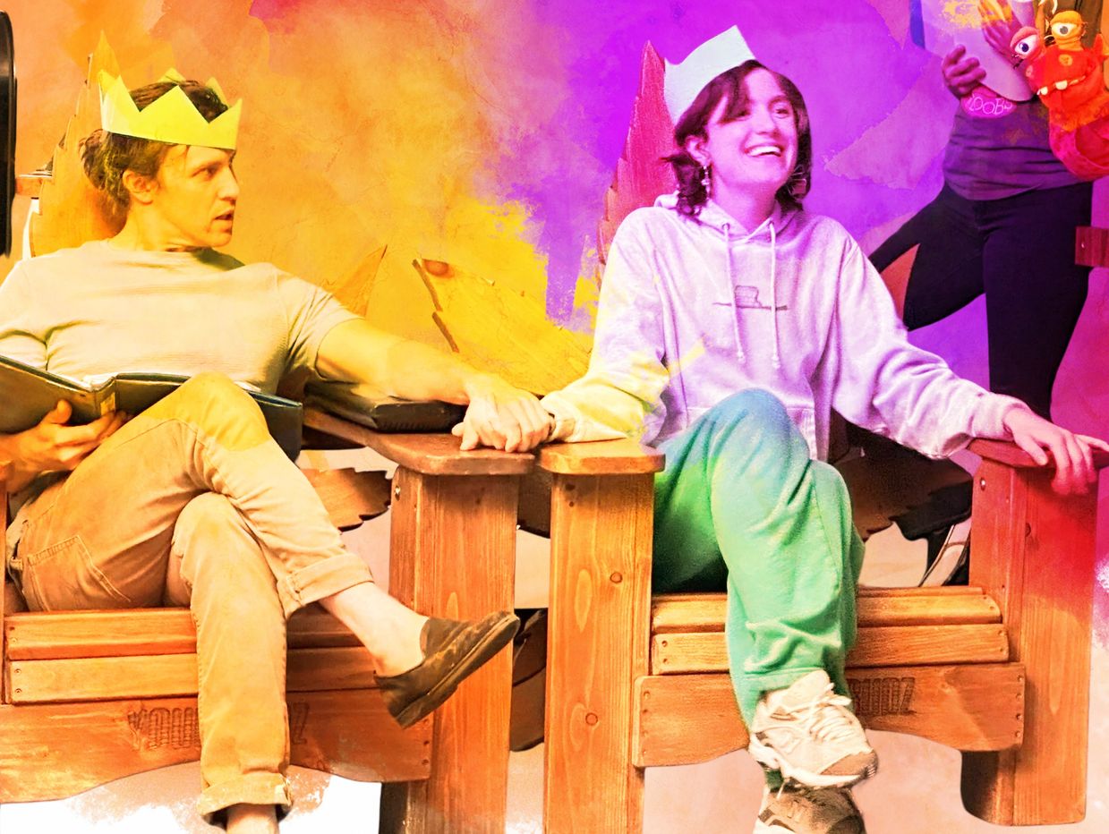 Two people wearing paper crowns holding hands while sitting on wooden thrones.